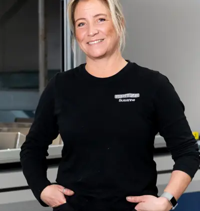 En kvinna som står vänd framåt. Hon är klädd i en svart tröja och står med händerna i fickorna och ser glad ut.